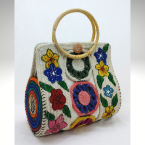 Handcrafted natural jute handbag with a round yellow bamboo-style handle, featuring intricate white rope floral embroidery on the front and a vibrant multicolor rope accent on the side.