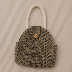 Handcrafted natural jute handbag with black woven pattern, rounded structured body, soft cream handles, button flap closure, fabric-lined interior, and earthy textured finish.