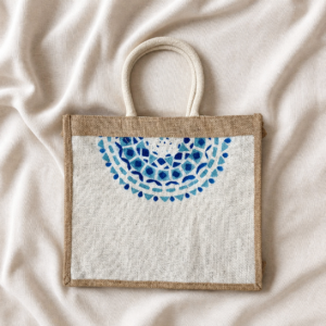 Front view of a handcrafted rectangular natural jute utility tote bag featuring padded cream handles, neutral jute trim, and a minimal blue artisan-inspired print on the upper front panel.